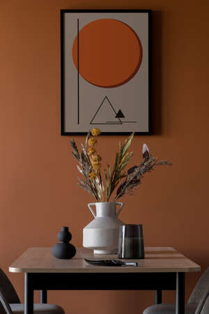 Stylish decorations, vase with flowers and candle on wooden dining table under modern poster on orange wallの写真素材