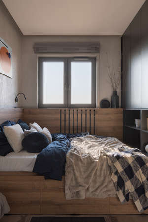 Small and cozy bedroom with window, comfortable bed in wooden frame with stylish bedclothes and many decorationsの写真素材