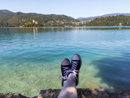 A traveler with black and white shoes sitting by clear and turquoise Bled lake with a view of the Church of the Assumption of Maria, Sloveniaの写真素材
