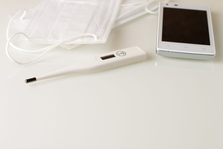 a white digital thermometer, a white mobile phone and a fragment of protective face masks lying on a...