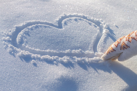 female hand in a knitted mitten drawing heart on white sparkling snow, St.Valentines Day romantic creative concept, sun shining, bright sunny winter day, snowy background, snowflakes, space for textの写真素材