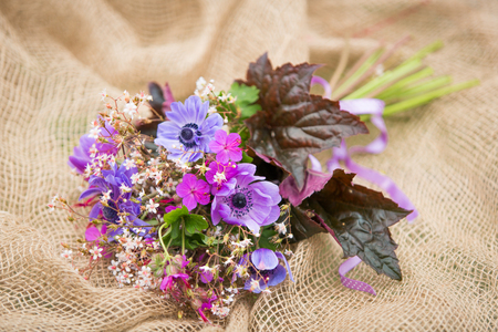 Garden style summer bouquet. Mix of lilac and purple anemone flowers, geranium flowers and heuchera or alumroot leaves. Bouquet on sackcloth background. Garden style wedding bouquet. Flower design.の写真素材