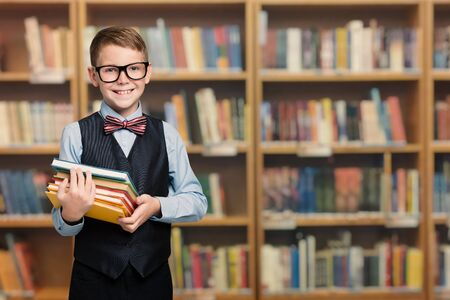 Happy Child in School Library Holding Books, Well Dressed Pupil Boy Portrait, Smiling Kid in Glasses over Bookshelvesの写真素材