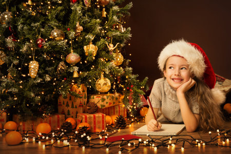 Christmas Kid Write Letter to Santa Claus. Child in Christmas Hat wishing New Year Gift on Xmas Eve in Holiday Room with Christmas Treeの写真素材