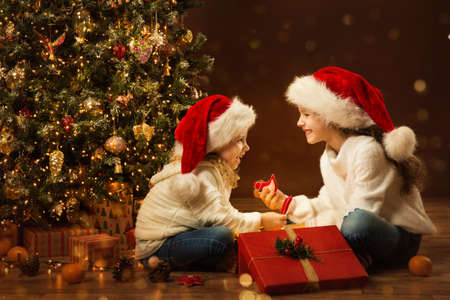 Christmas Child Girl opening Box and giving Gift Toy to Sister. Happy Children in Santa Hat next to Xmas Tree. Fairy Magic Interior, Indoor Lightsの写真素材
