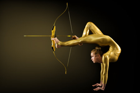 Archer Shooting by Legs with Gold Bow and Arrow. Flexible Gymnast aiming Target standing on Hand upside down. Goal Achievement Concept, Studio shot over Black backgroundの写真素材