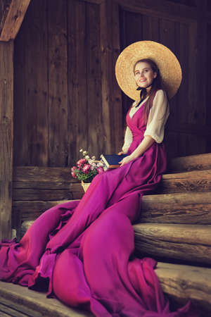 Romantic Vintage Woman in Summer Hat dreaming with Book on wooden Stairs Background. Victorian Fashion Portraitの写真素材
