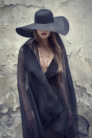 High Fashion Model in Black Hat hidden Face Grunge Wall Background. Stylish Woman Portrait with Red Lips and Necklace Jewelry. Elegant Retro Ladyの写真素材
