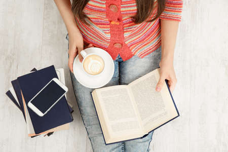 Close up Women Hands Holding Coffee Cup and Reading opened Book. Top view of Woman with Teacup having Break. Sitting Young Girl Study Booksの写真素材