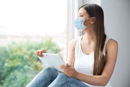 Woman in Medical Mask isolated Stay Home. Girl in Facemask Use Tablet Working at Window Sill. Self Isolation and Distance Studyの写真素材