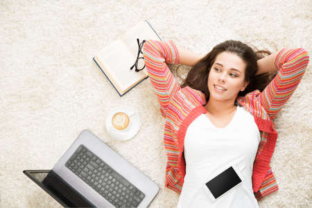 Happy Woman Relax at Home with Laptop and Phone. Young Girl have Coffee Break. Student Lying Down Hands behind Head on White Carpet Floorの写真素材