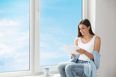 Woman using Tablet sitting on Window Sill at Home. Young Girl Reading White Digital Touch Pad PC and drinking Coffee. Window Glass with Blue Sky Backgroundの写真素材