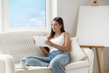 Woman reading Tablet PC sitting on Sofa in front of empty Easel Canvas. Young Girl using Touch Pad Screen resting next to Window. Woman with Tablet at Homeの写真素材