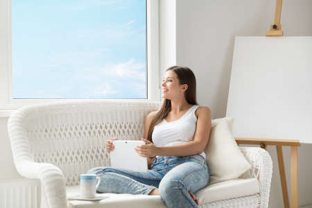 Woman with Tablet Thinking Looking forward at Home Sofa. Beautiful Girl Dreaming with Touch Pad next to Window and Drawing Easel Canvas. Woman planning looking At Skyの写真素材