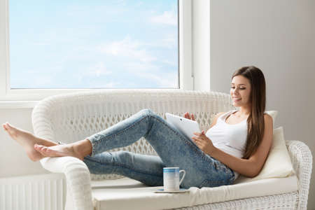 Woman reading Tablet at Home lying down at Sofa next Window. Morning Coffee Break. Beautiful Happy Girl resting with Digital Touch Pad in White Roomの写真素材