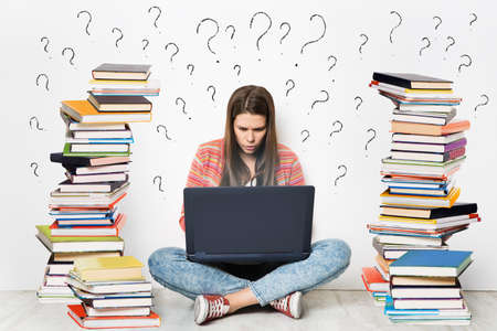 Woman using Laptop. Student Girl working on Computer Search in Internet around Stacks of Books. Question Marks Drawn On White Wall Background. E Learning, Online Studying, Reading Books in Digital Libraryの写真素材