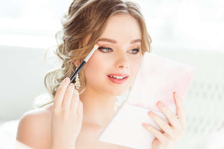 Bride Wedding Makeup. Bridal Make up. Beautiful Woman looking at Hand Mirror and putting Eye Shadow Eyebrow Self Make up in White Roomの写真素材
