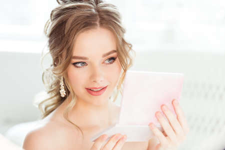 Beautiful Bride Make up Portrait. Bridal Wavy Hairstyle. Wedding Professional Makeup. Beauty Woman Model looking at Hand Mirror in Sunny Bright Salon Roomの写真素材
