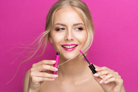 Blonde Model Girl holding Pink Lipstick Brush over Bright Magenta Background. Summer Beauty Woman Face Cosmetics Make up. Fashion Model applying Lip Gloss Makeupの写真素材