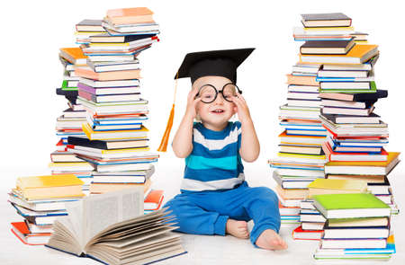 Little Baby Boy Reading Books. Happy Smiling Child in Graduation Cap Studying. Funny Kid wearing Glasses sitting next to Stack of Books over White Backgroundの写真素材