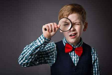 Little Kid Looking Through Magnifying Glass Lens. Funny Surprised Child Holding Loupe. School Boy in Smart Casual Clothing over Gray Studio Backgroundの写真素材