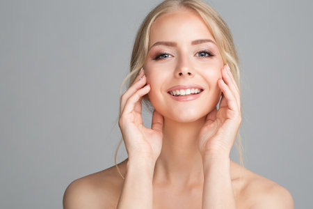Cheerful Smiling Young Woman Massaging Face Cheek Bones. Beauty Model with Perfect Smooth Skin looking at Camera over Gray Backgroundの写真素材