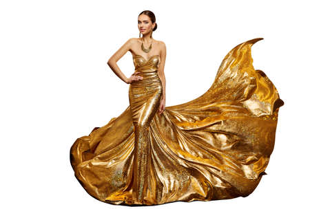 Woman in Long Golden Evening Dress waving in Air. Elegant Fashion Model in Shiny Luxury Gown with Gold Jewelry over Studio White Backgroundの写真素材