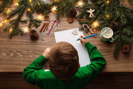 Child Writing Christmas Letter to Santa Claus. Top View of Kid sitting at Wooden Table with Xmas Tree Decoration Lights and Empty White Paper Wish Listの写真素材
