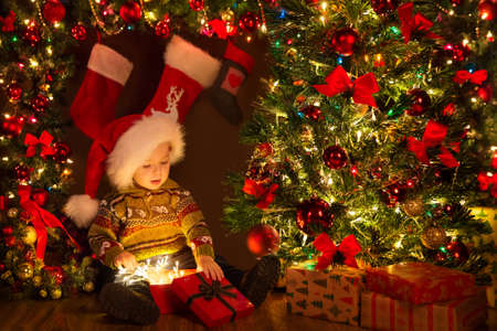 Christmas Kid opening Gift Box with Xmas Lights Garland. Cute Baby in Santa Hat sitting next to Decorated Fir Tree in Dark Room Interior with Shining Ornamentsの写真素材