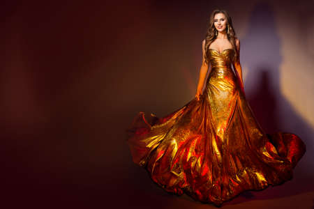 Fashion Model in Long Golden Dress. Smiling Beauty Woman in Evening Shining Fluttering Gold Gown walking over Dark Brown Backgroundの写真素材