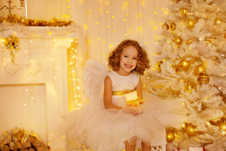 Little Girl Holding Candle sitting next to Christmas Tree Decorated with Golden Ornaments and Lights. Happy Child in White Room Interior with New Year Decorationsの写真素材