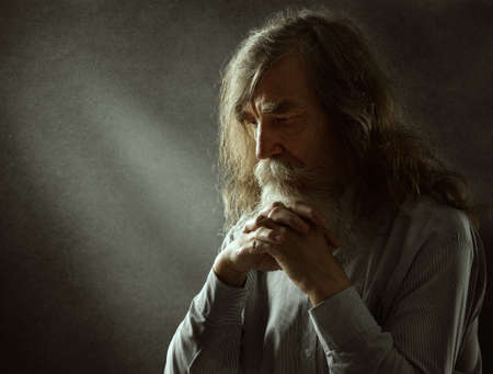 Old Man Praying. Sad Thinking Senior with White Beard over Dark Background with Light Beams. Elder Men Worship Face Portrait with Palms togetherの写真素材