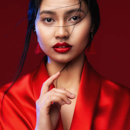 Asian Beauty Woman with Red Lips Make up. Chinese Beautiful Model Face Portrait over Red Background. Fashion Japanese Girl looking at camera in Silk Kimono close upの写真素材