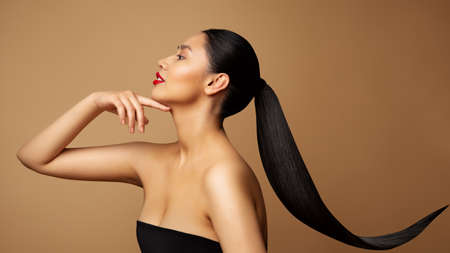 Beauty Model Profile. Young Woman with long Ponytail Hair. Women Face Side view over beige background. Lady with Red Lipstick and Black Straight Tail Hairstyleの写真素材