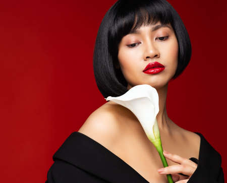 Fashion Model with Red Lipstick. Asian Woman Beauty with Black Bob Hairstyle over Red Background. Elegant Lady Face Portrait with White Calla Lily Flower. Stylish Women Short Haircut. Chinese Beautiful Girlの写真素材