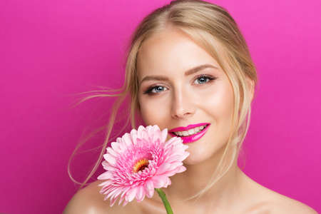 Beauty Model with Pink Lipstick. Smiling Young Woman with Smooth Skin Makeup and Lip Gloss. Fashion Girl Portrait with Glossy Lipstick with Flower over Purple Background. Women Cosmetics and Summer Facial Careの写真素材