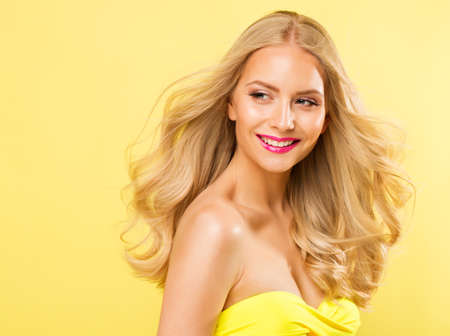 Blond Woman with Long Curly Hairstyle. Blonde Fashion Girl with Summer Face Makeup enjoying Sun. Beauty Model Happy smiling Portrait over bright Studio backgroundの写真素材