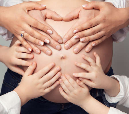 Pregnant Belly with Parents and Kids Hands. Father Heart shaped Hands and little Children touching Mother Pregnancy Abdomen. Big United Family Love and Child Birth Conceptの写真素材