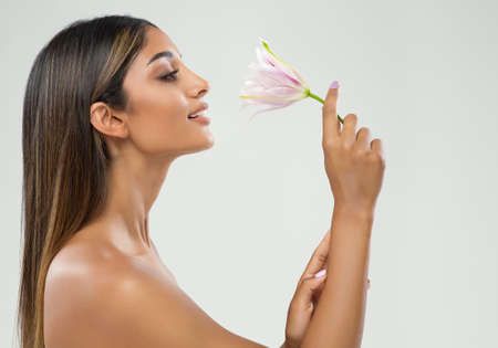 Beauty Spa Face Model Profile over White. Young Woman with tanned Skin Smelling Lily Flower. Beautiful Brunette Girl with Long Straight Hair happy smiling Side View over Grayの写真素材