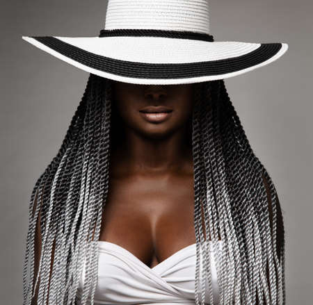 African Woman with Long Braids Hair. Black and White Concept. Beauty Model in Big Hat hidden Face. Afro Hairstyle and Lips Makeup. Sexy Mysterious Women Portrait over Gray Studio Backgroundの写真素材
