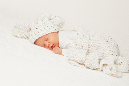 Baby sleeping with Blanket. Newborn lying on Stomach in White Bed. Cute Baby Face Portrait in Knitted Hat. One Month Child Bedtimeの写真素材