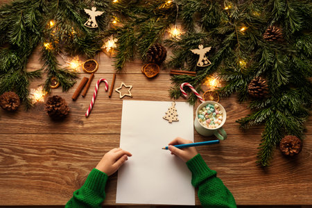 Letter to Santa. Christmas Child Hands Close up writing Xmas Gifts Shopping Wish List over brown Wooden Table. Wood Background with Fir Tree Branch decorated Christmas Lights and Toysの写真素材