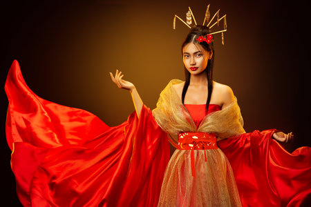 Asian Empress in Red Silk Dress with Golden Jewelry. Beautiful Chinese Girl in Traditional Wedding Costume Clothes. Japanese Model in Fantasy Bride Gown over Dark Studio Backgroundの写真素材