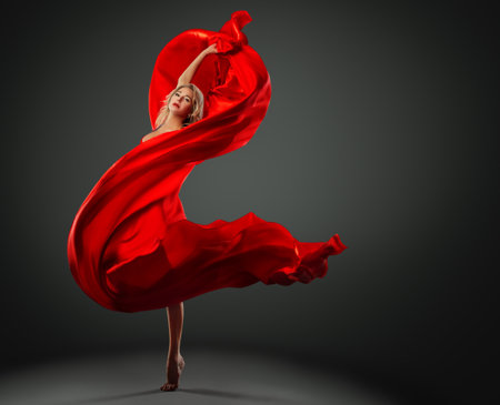 Ballerina dancing with Red Silk Fabric flying on Wind. Modern Ballet Dancer jumping over Dark Studio background. Fashion Woman in Red Dress wrapped in waving Scarfの写真素材