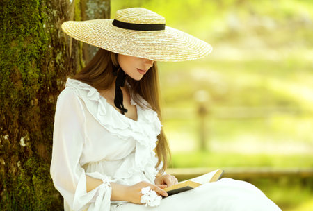 Romantic Girl in White Dress and Straw Sun Hat reading Book sitting under Tree. Young Woman studying in Spring Park. Old Fashioned Elegant Lady outsideの写真素材