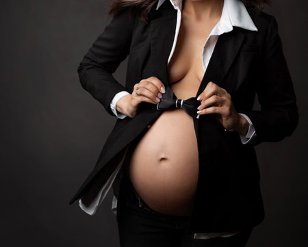 Close up of Pregnant Woman Belly with Black Bow Tie over Dark Gray Background. Elegant Future Mother in Classic Business Suit waiting Baby Boy. Pregnancy Fashion Style. Gender Identity Conceptの写真素材