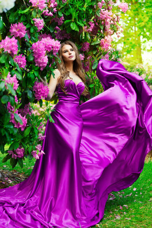Beautiful Dreamy Woman in long Purple Dress in Blossom Garden. Fantasy Girl in Spring Park enjoying Flower Smell. Fashion Model in Satin Pink Gown with Outdoorsの写真素材