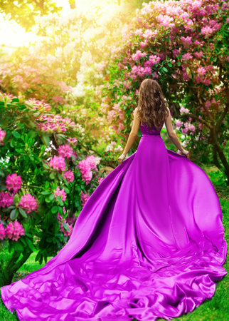 Beautiful Woman in long Purple Dress in Flower Garden Rear View. Fantasy Girl walking away in Spring Blossom Park enjoying Sun Light. Fashion Model in Satin Pink Gown with Train Outdoorsの写真素材