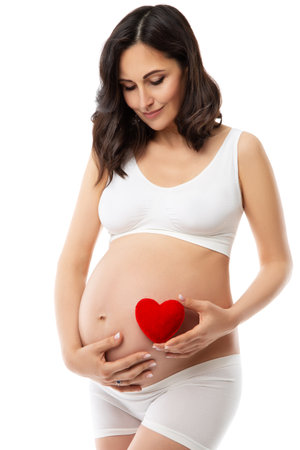 Happy Pregnant Woman holding Red Heart on Belly. Pregnancy Health Body Skin Care. Beautiful Future Mother in White Cotton Underwear Clothes. Maternity Healthcare and Cosmetologyの写真素材