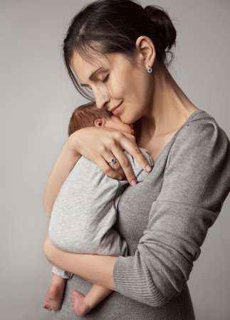 Happy Mother holding Baby in Arms. Newborn Health Care and Development. Beautiful Mom embracing little Son over Gray Background. Smiling Woman enjoying parentingの写真素材